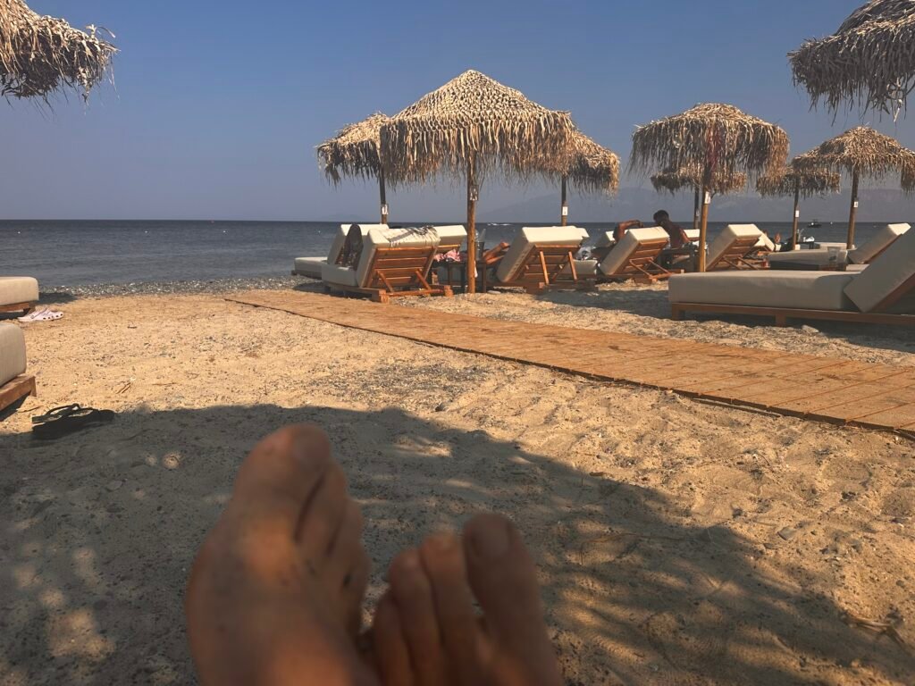 Uitzicht vanaf een ligbed op het strand: blote voeten in het zand, daarachter strandbedden met rieten parasols, de zee en bergen in de verte.